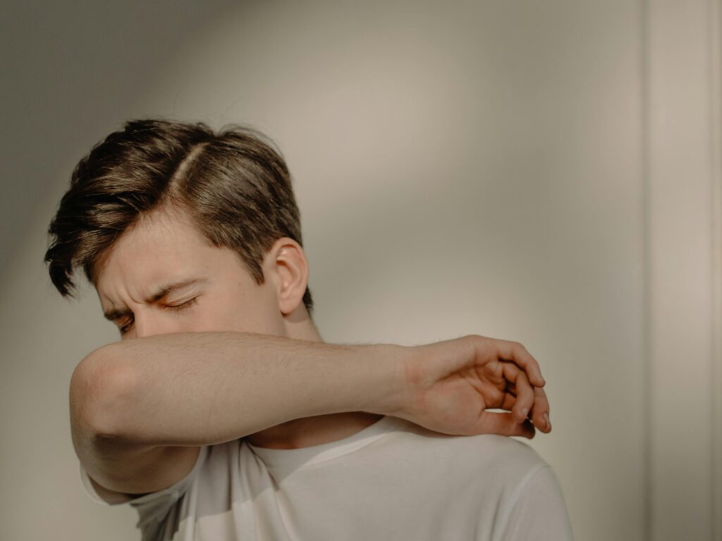 A young man sneezes into his elbow, showcasing a hygiene practice, in an indoor setting.