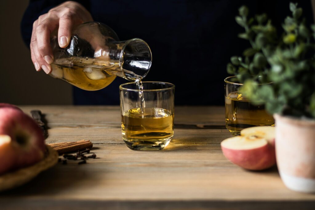 Close-up of someone pouring apple cider into glasses on a wooden table with apples and spices.